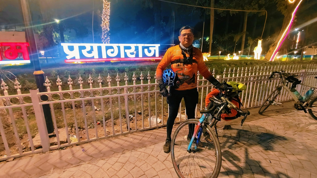 A cyclist stands next to a brightly lit 'प्रयागराज' sign at night, with bicycles parked nearby.